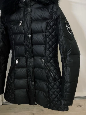 Svart dunjacka med päls - Snygg svart dunjacka från Rockandblue med figurnära passform, quiltade detaljer och svart päls på huvan. Jackan har flera dragkedjeförsedda fickor,Perfekt för kalla vinterdagar.lite slitage på armbågen men syns knappast 