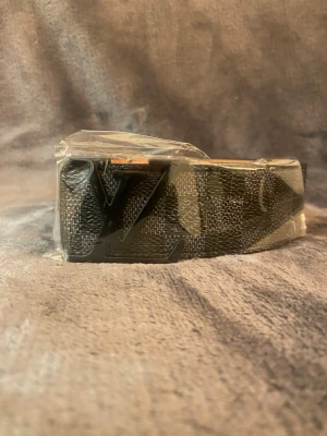 Svart LV-bälte med monogramspänne - Snyggt svart bälte från Louis Vuitton med klassiskt LV-monogram på metallspännet. Bältet har diskret mönster i tyget och insidan är beige. Perfekt accessoar för att lyfta din outfit.