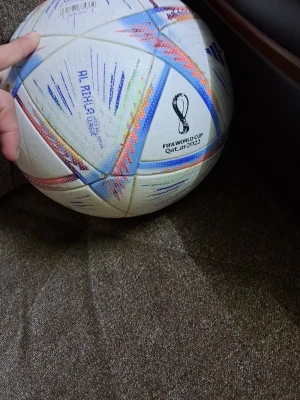 Vit fotboll Adidas Al Rihla - Säljer en vit fotboll av modellen Al Rihla från Adidas. Bollen har färgglada detaljer i blått, orange och lila samt tryckt logga. Tillverkad i syntetmaterial och har klassisk paneldesign. Perfekt för fotbollsträning eller spontanspel med vänner.