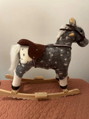 Gunghäst i grått och vitt med sadel - Mysig gunghäst i grå och vit plysch med bruna detaljer och sadel. Hästen har vita prickar, mjuk man och svans samt trämedar. Perfekt för lek och inredning i barnrummet.