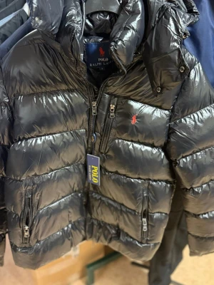 Svart pufferjacka från Polo Ralph Lauren - Snygg svart pufferjacka från Polo Ralph Lauren med glansig finish, huva och dragkedja framtill. Jackan har två sidofickor med dragkedja och den klassiska röda loggan broderad på bröstet. Perfekt för kalla vinterdagar och riktigt trendig look.