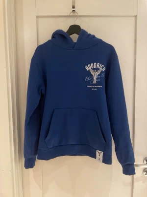 Blå hoodie från Hoodrich  - Blå hoodie från Hoodrich med vit text och tryck på ryggen. Tryck även på bröstet och huvan. Klassisk känguruficka framtill och mjukt material, perfekt för en streetwear-look. Modellen är 185 cm.