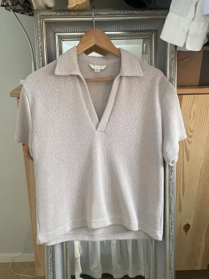 Ljusbeige stickad polotopp S - Ljusbeige stickad polotopp med korta ärmar och djup v-ringning. Toppen har en avslappnad passform och mjukt material som känns skönt mot huden. Perfekt till jeans eller kjol för en enkel men snygg look.