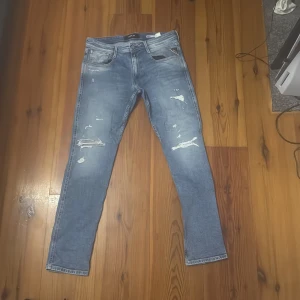 Blå slitna slim fit-jeans Replay - Säljer ett par blå slim fit-jeans från Replay med slitningar och hål på framsidan. Jeansen är i storleken W32 och L32. Jeansen är inte alls mycket används och är i bra skick. Modellen är Anbass.