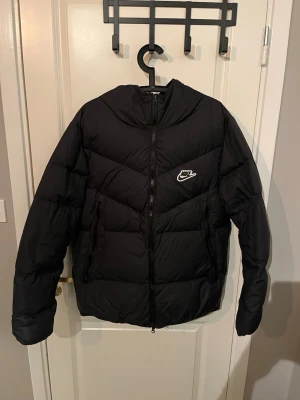 Svart dunjacka från Nike med huva - Snygg svart dunjacka från Nike med dragkedja framtill och två sidofickor. Jackan har en avtagbar huva och en vit Nike-logga på bröstet. Puffig modell som håller värmen under kalla dagar. Perfekt för vintern och riktigt stilren. Bra skick om man bortser de från dom sista bilderna. 