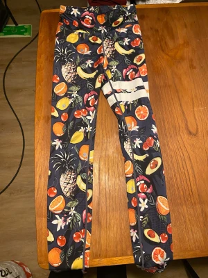 Mörkblå tights med fruktmönster - Snygga mörkblå träningstights från Stronger med färgglatt fruktmönster som ananas, avokado, apelsin, körsbär och banan. Tightsen har hög midja och är tillverkade i stretchigt funktionsmaterial som passar perfekt till gymmet eller löpning. Loggan har börjat att försvinna (se bild).