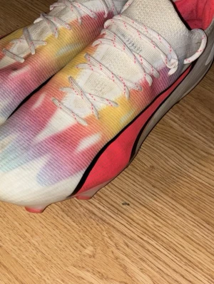  Puma fotbollsskor - Säljer ett par snygga Puma fotbollsskor med coolt färgskiftande mönster i vitt, rött, gult och blått. Skorna har klassisk snörning, låg profil och är tillverkade i syntetmaterial för bästa känsla på planen. Perfekta för dig som vill sticka ut på matchen!
