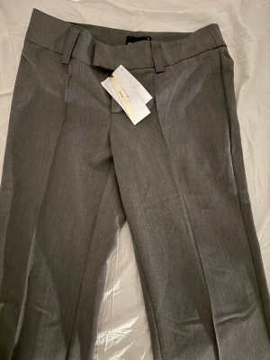 Grå kostymbyxor med raka ben - Snygga grå kostymbyxor med raka ben och låg midia . Byxorna har breda hällor för bälte och två fickor med lock bak. Materialet ser ut att vara en blandning av ull och polyester för en klassisk look.(aldrig använt köpte i för liten storlek)