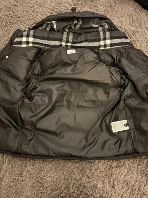 Svart dunjacka från Burberry - Snygg svart dunjacka från Burberry med klassiskt rutigt foder i grått, vitt och svart. Jackan har en quiltad insida och är tillverkad i polyester med dunfyllning. Perfekt för kalla dagar och har en tidlös design med lyxig känsla.