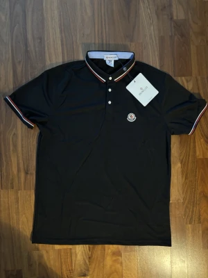 Svart Moncler pikétröja med ränder - Svart pikétröja från Moncler med klassisk krage och knappar framtill. Kragen och ärmsluten har snygga ränder i vitt och rött. Moncler-logga på bröstet. Perfekt för en stilren och sportig look.