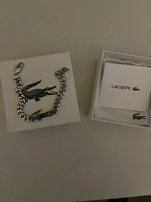 Silverfärgat armband från Lacoste - Snyggt silverfärgat armband från Lacoste med en kedjedesign och den klassiska gröna krokodilen som detalj. Armbandet har ett robust spänne och kommer i originalask med Lacoste-logga. Perfekt accessoar för dig som gillar stilrena smycken med ikonisk touch. Perfekt som present till alla hjärtans dag.