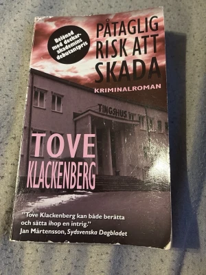 Påtaglig risk att skada av Klackenberg - Kriminalroman av Tove Klackenberg i pocketformat med omslag i svart, vitt och rosa. Framsidan visar ett tingshus och boken bjuder på en spännande berättelse om rättsprocesser och intriger i Värmland. Perfekt för dig som gillar deckare och thrillers.