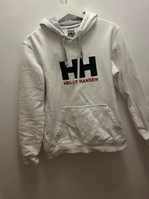 Vit hoodie från Helly Hansen - Snygg vit hoodie från Helly Hansen med stor svart HH-logga och röd text på bröstet. Hoodien har en klassisk känguruficka, grå dragsnören och mjukt material på insidan. Perfekt för en chill och avslappnad stil.
