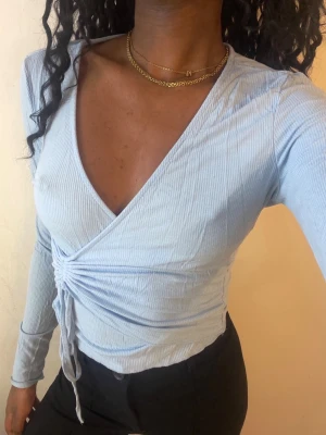 Ljusblå omlottopp med snörning - Trendig ljusblå omlottopp med ribbad struktur och v-ringning. Toppen har långa ärmar och snörning i sidan som ger en snygg detalj och justerbar passform. Perfekt att styla med både jeans och kjol.
