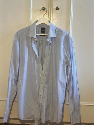 Blåvit randig skjorta från W Collection - Klassisk blåvit randig skjorta från Oscar Jacobsson med lång ärm och knappar framtill. Skjortan har en avslappnad krage och är tillverkad i ett lätt bomullstyg, perfekt för en stilren look.