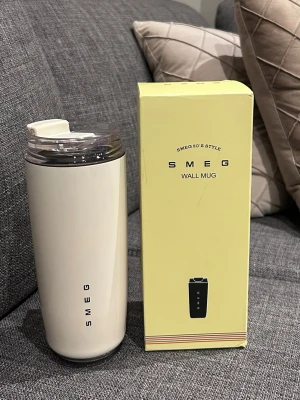 Smeg Wall Mug beige med lock - Snygg beige Wall Mug från Smeg i retro 50-talsstil. Muggen har ett genomskinligt plastlock med öppning för att dricka och Smeg-logga på sidan. Perfekt för kaffe eller te på språng. Levereras i originalförpackning.