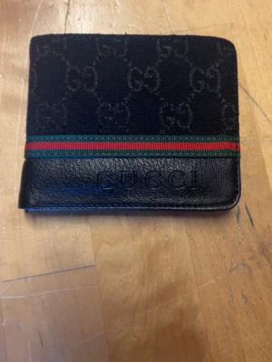Svart Gucci plånbok med GG-mönster - Snygg svart plånbok från Gucci i läder och canvas med klassiskt GG-mönster och grön-röd rand. Plånboken har flera kortfack och präglad Gucci-logga både på utsidan och insidan. Perfekt för dig som vill ha en stilren och lyxig accessoar.