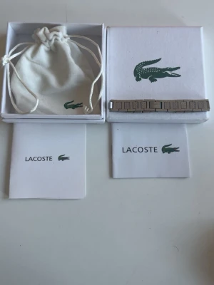 Silverfärgat armband från Lacoste - Snyggt silverfärgat armband från Lacoste i metall. Kommer i original ask och tygpåse med klassiska Lacoste-krokodilen. Perfekt accessoar för dig som gillar stilrena detaljer, priset kan diskuteras. Helt ny bara boxen som ser lite sliten ut.