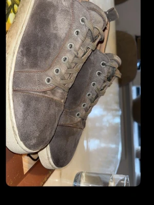 Bruna mockasneakers med röd sula - Snygga sneakers i grå mocka med vita kanter och en unik röd sula. Skorna har klassisk snörning och en stilren design som passar till det mesta. Perfekta för dig som gillar en clean look med en twist. Ovandelen är i mjukt mockamaterial. Kvitto och box finns med.