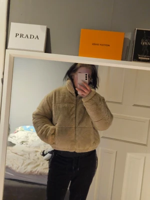 Beige teddyjacka med hög krage - Mysig beige teddyjacka med hög krage och dragkedja framtill. Jackan har en boxig passform och två sidofickor. Perfekt för kalla dagar när du vill vara både varm och trendig. Materialet är fluffigt och ger en cool streetstyle-look.