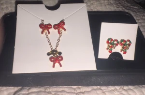 Ett julhalsband med julklapps rosetter, julörhängen med julklapps rosetter  - Säljer ett par örhängen formade som polkagrisar i rött, grönt och vitt med små guldfärgade rosetter. Perfekta för dig som vill ha en färgglad och lekfull julstil. Örhängena är tillverkade i metall med emaljerade detaljer.