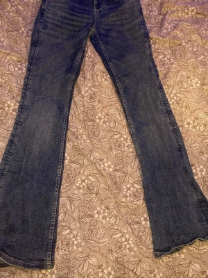 Blå bootcut jeans med detaljer på bakfickorna - Säljer ett par klassiska blå bootcut jeans i denim med lätt slitna detaljer och femficksmodell. Jeansen har normal midja och utsvängda ben, perfekta för en avslappnad och trendig look. Passar dig som gillar bootcut och vill ha ett par bekväma jeans.