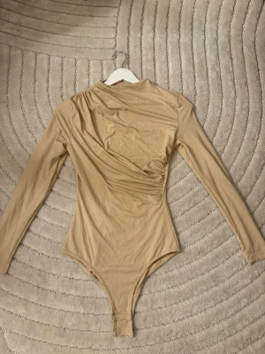Beige långärmad body - Säljer en beige body med långa ärmar och rund hals. Bodyn har en åtsittande passform och är tillverkad i ett stretchigt, mjukt material som ger en smickrande siluett. Perfekt att styla med jeans eller kjol för en clean look.