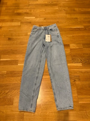 Ljusblå raka jeans från Vero Moda modell ”Tessa mom” - Säljer ett par ljusblå jeans från Vero Moda med rak passform och klassisk femficksdesign. Jeansen har hög midja, bälteshällor och stängs med knapp och dragkedja. Materialet är 100% bomull för en skön och avslappnad känsla. STORLEK:26x32! 