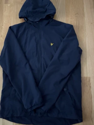 Mörkblå vindjacka från Lyle & Scott - Snygg mörkblå vindjacka från Lyle & Scott med huva och dragkedja framtill. Jackan har det klassiska gula logomärket på bröstet och två sidofickor. Perfekt för blåsiga dagar och enkel att matcha med olika outfits.