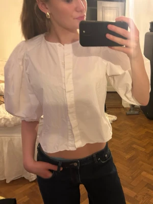 Vit puffärmad blus med knappar - Snygg vit blus med puffiga korta ärmar och knäppning framtill. Blusen har en rak, något croppad passform och är tillverkad i ett lätt bomullstyg. Perfekt för en trendig och stilren look.