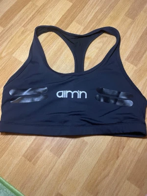 Svart sport-bh från Aim'n - Snygg svart sport-bh från Aim'n med brottarrygg och vita logotypdetaljer framtill. Bh:n har elastiskt material och dekorativa svarta ränder i glansigt tryck både fram och bak. Perfekt för träning och ger bra stöd. HELT NY!!!! Utan lapp.