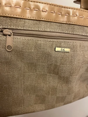 Beige handväska från BG med detaljer - Snygg beige handväska från BG med rutmönstrad textil och ljusbruna detaljer i konstläder. Väskan har ett ytterfack med dragkedja och två rejäla handtag. Perfekt för dig som gillar stilrena accessoarer med en twist.