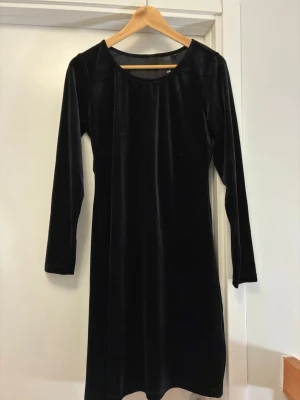 Svart sammetsklänning med lång ärm - En svart klänning i mjukt sammetsliknande material med lång ärm och rund halsringning. Klänningen har en rak och enkel passform med en guldfärgad dragkedja i ryggen som en fin detalj.