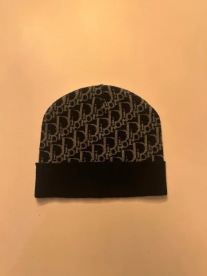Svart Dior beanie med logomönster - Snygg svart beanie från Dior med grått logomönster över hela mössan. Klassisk uppvikt kant och mjukt material som sitter skönt på huvudet. Perfekt accessoar för dig som vill ha en trendig och exklusiv look.