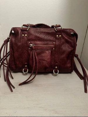 Vinröd handväska med flätade handtag - Snygg vinröd handväska i fejkskinn med flätade handtag och dekorativa dragkedjor. Väskan har detaljer i guldfärg och långa tofsar på sidorna för en cool look. Perfekt storlek för det viktigaste och ger en edgy vibe till din outfit. Helt oanvänd.