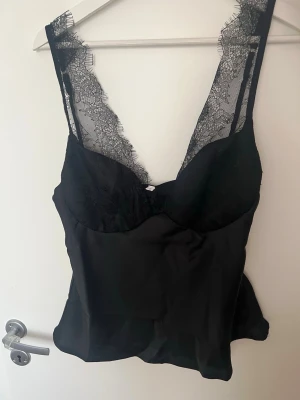 Svart spetslinnetopp med v-ringning - Snygg svart linnetopp med djup v-ringning och spetsdetaljer längs axelbanden och urringningen. Toppen har en slät, glansig yta och tunna axelband i spets som ger en elegant look. Perfekt att styla med jeans eller kjol.