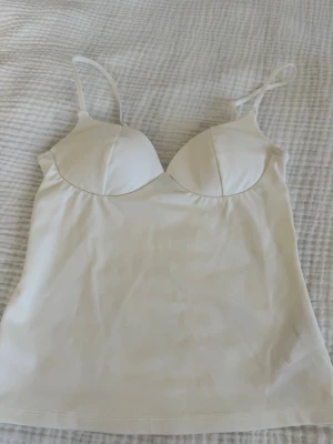 Vit body med tunna axelband - Säljer en stilren vit body med tunna justerbara axelband och vadderade kupor. Materialet är mjukt och stretchigt, perfekt för att ge en smickrande passform. Enkel och clean design utan synliga detaljer eller mönster.