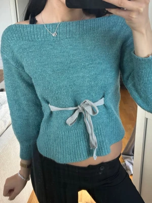 Turkos stickad tröja med rosett - Mysig turkos stickad tröja med bred ribbad kant och en grå rosett i midjan. Tröjan har en off shoulder-design och långa ärmar. Perfekt för dig som vill ha en trendig och bekväm look.