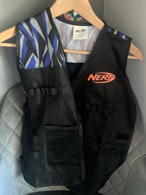 Svart Nerf-väst med blått mönster - Cool svart väst från Nerf med blått och grått grafiskt mönster upptill. Västen har flera elastiska band och fickor för tillbehör, perfekt för lek och action. Nerf-loggan i orange framtill ger en sportig vibe.