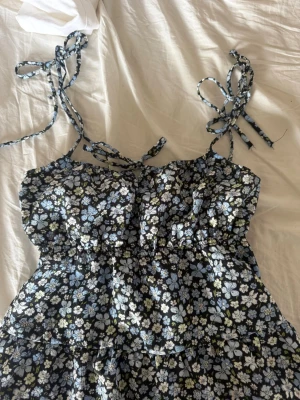 Blommig ärmlös klänning med knytband - Supersöt klänning med svart botten och blommigt mönster i blått, vitt och grönt. Ärmlös modell med smala axelband som knyts på axlarna för en personlig touch. Klänningen har en lätt rynkad midja och luftig passform, perfekt för varma dagar.