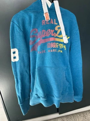 Blå Superdry hoodie med tryck - Snygg blå hoodie från Superdry med färgglatt tryck på bröstet och japanska tecken. Stor vit siffra 82 på ena ärmen, huva med vita snören och känguruficka framtill. Perfekt för en avslappnad streetstyle-look.