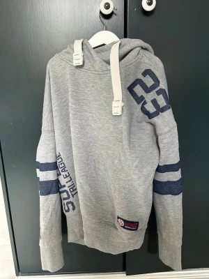 Grå hoodie med blå tryck och huva - Snygg grå hoodie från Superdry med blå tryck på ärmar och axel, samt breda blå ränder runt armarna. Stor huva med vita, grova dragsnören och ribbade muddar. Perfekt för en avslappnad streetstil.