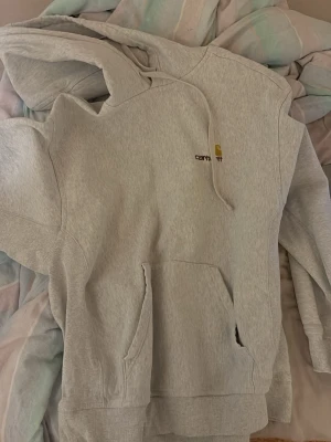 Ljusgrå hoodie från Carhartt - Ljusgrå hoodie från Carhartt med broderad logga på bröstet. Klassisk känguruficka framtill och justerbar huva med dragsko. Mjuk bomullsblandning och ribbade muddar vid ärmslut och nederkant. Perfekt för en avslappnad streetwear-look.