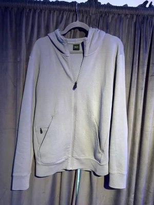 Ljusgrå hoodie från BOSS - Snygg ljusgrå hoodie från BOSS med dragkedja framtill, två fickor med dragkedja och diskret logga på bröstet. Klassisk huva och ribbade muddar. Perfekt för en avslappnad och stilren look.