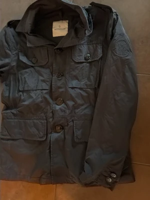 Svart jacka från Moncler med huva - Stilren svart jacka från Moncler med huva och flera fickor framtill. Jackan har knäppning med stora knappar och en diskret Moncler-logga på ärmen. Perfekt för dig som gillar klassisk och funktionell design.