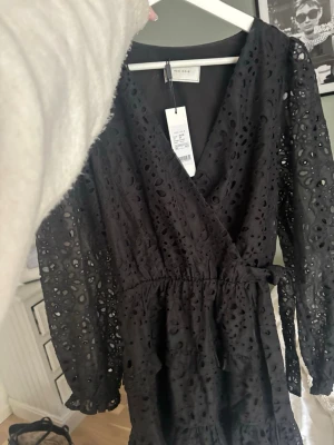 Svart omlottklänning från Neo Noir - Svart omlottklänning från Neo Noir med broderade hål och volangdetaljer. Klänningen har v-ringning, långa ärmar och knyt i midjan. Tillverkad i bomull och har en kort längd som ger en trendig look.