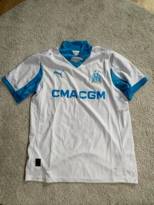 Marseille matchtröja Puma vit/blå - Snygg vit matchtröja från Olympique de Marseille med blå detaljer och logga på bröstet. Puma-logga och sponsortryck framtill. Korta ärmar och sportig passform, perfekt för fotboll eller träning.