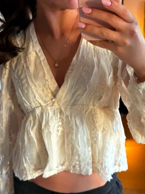 Vit blus med spetsdetaljer och v-ringning - Romantisk vit blus med v-ringning och transparenta spetsdetaljer. Blusen har lång ärm och volang nedtill för en luftig känsla. Perfekt för dig som gillar en feminin och trendig stil. Materialet är tunt och lätt, vilket ger en elegant look.