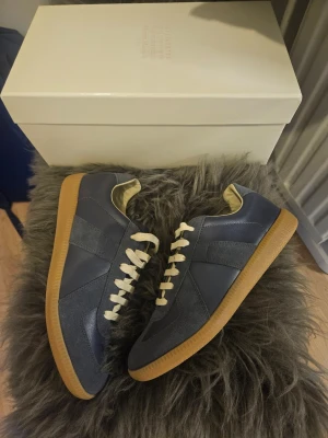 Maison Margiela Pewter GråBlå Gats - Snygga sneakers från Maison Margiela i mörkblått läder och mocka med vita skosnören och klassisk gummisula. Modellen har en retroinspirerad design och är perfekta för dig som gillar stilrena och tidlösa sneakers.