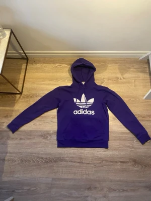 Lila Adidas hoodie med tryck - Snygg lila hoodie från Adidas med den klassiska vita trefoil-loggan och Adidas-tryck på bröstet. Hoodien har huva och en stor magficka framtill. Perfekt för dig som gillar streetwear och vill sticka ut med färg.